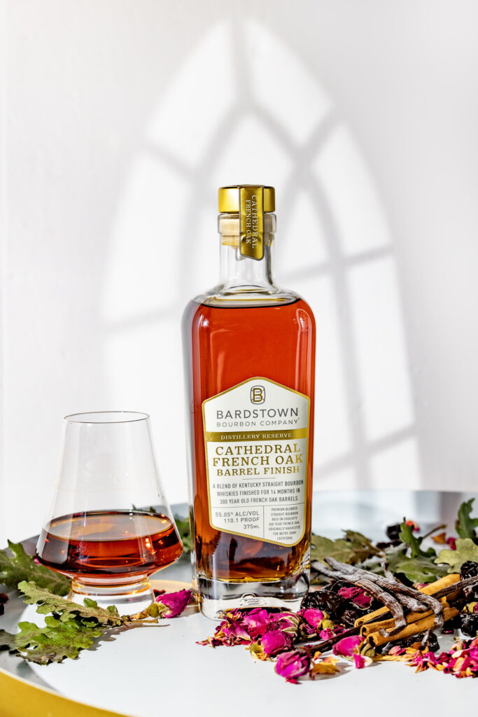 Bottle of Bardstown Bourbon Company’s Cathedral French Oak Barrel Finish with a glass of bourbon beside it, surrounded by dried rose petals, herbs, and spices on a white surface.