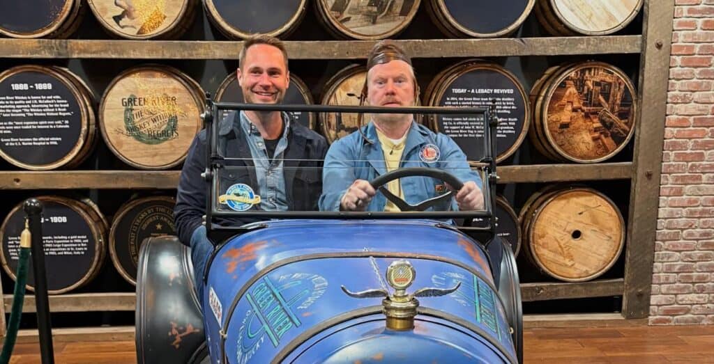Two men sitting in a vintage-style blue car in front of a wall of whiskey barrels. The man on the right is comedian Jim Gaffigan, playfully posing as the driver.