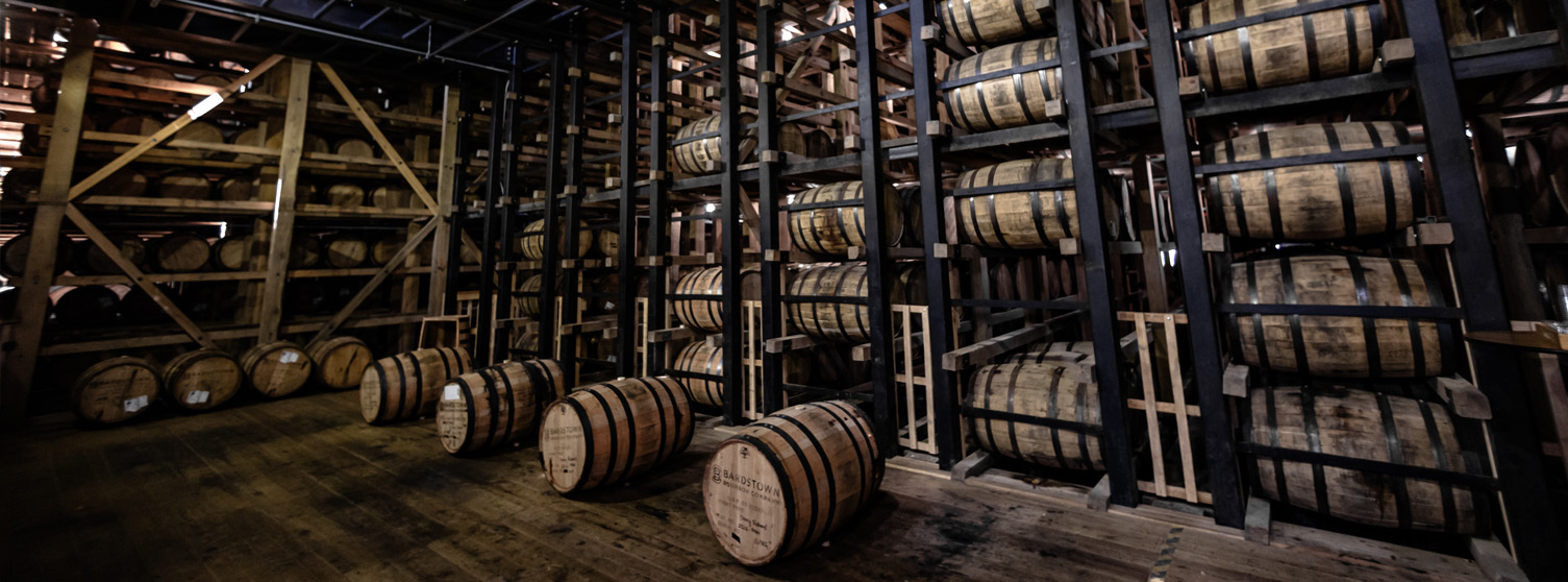 Interior of a rickhouse with rows of Bardstown Bourbon Co. barrels aging on wooden and metal racks, with several barrels on the floor.