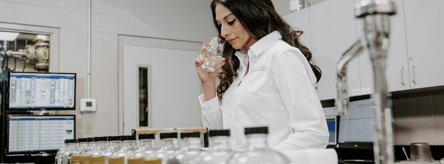 Woman in a Bardstown Bourbon Company lab coat evaluating a sample by smell in a lab setting, with rows of labeled bottles and computer monitors in the background.