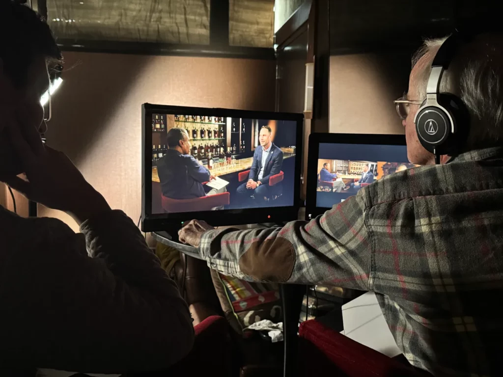 Behind-the-scenes view of a video production monitor showing two men in a bar setting having an interview. A crew member wearing headphones reviews the footage on screen.