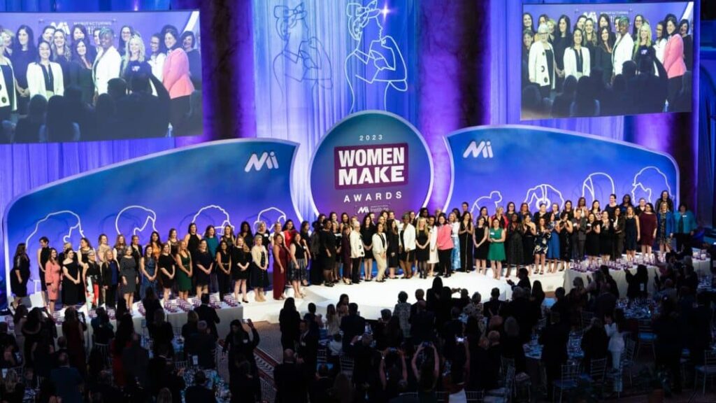 Large group of women standing on stage at the 2023 Women MAKE Awards ceremony, hosted by the Manufacturing Institute, with a cheering audience and purple-lit backdrop.